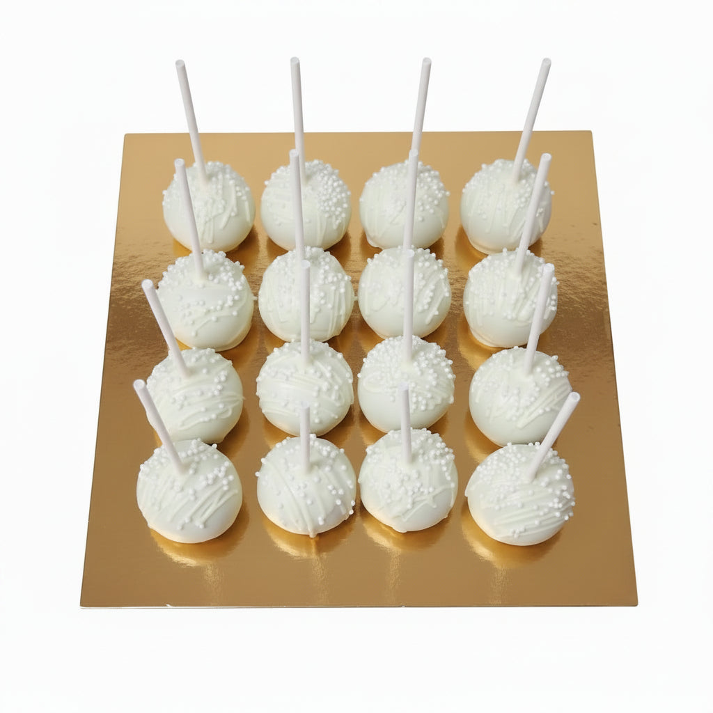 A cardboard box containing 12 white cake pops with white licks, placed on a green cutting mat on a dark table.