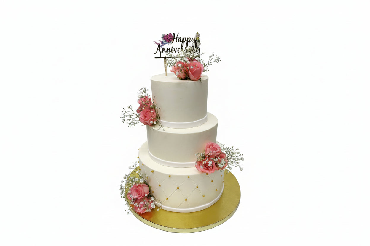 A multi-tiered white anniversary cake decorated with peach roses and a 'Happy Anniversary' topper, presented on a gold platter.