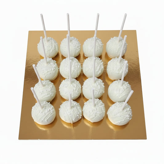 A cardboard box containing 12 white cake pops with white licks, placed on a green cutting mat on a dark table.