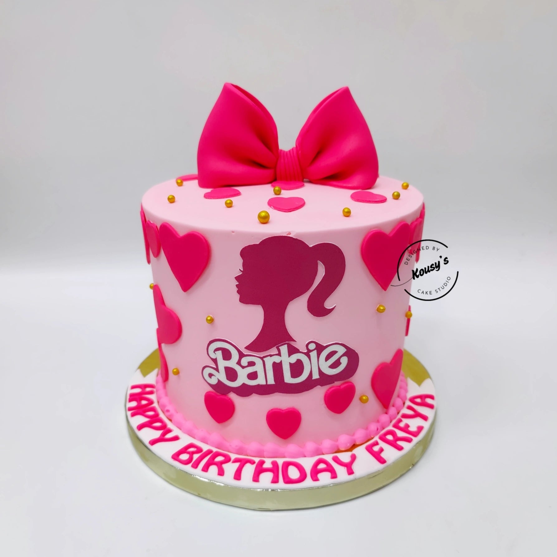 A pink Barbie-themed birthday cake with a large pink bow on top, pink heart decorations, gold sprinkles, and the Barbie logo on the front. The cake sits on a white board with 'HAPPY BIRTHDAY FREYA' written in pink icing, set against a plain light gray background.