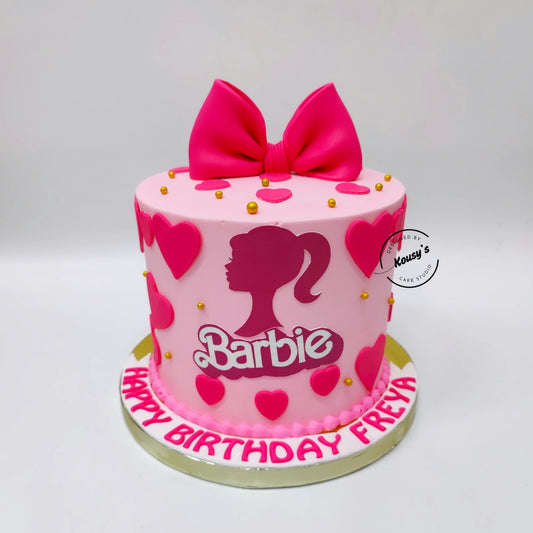 A pink Barbie-themed birthday cake with a large pink bow on top, pink heart decorations, gold sprinkles, and the Barbie logo on the front. The cake sits on a white board with 'HAPPY BIRTHDAY FREYA' written in pink icing, set against a plain light gray background.