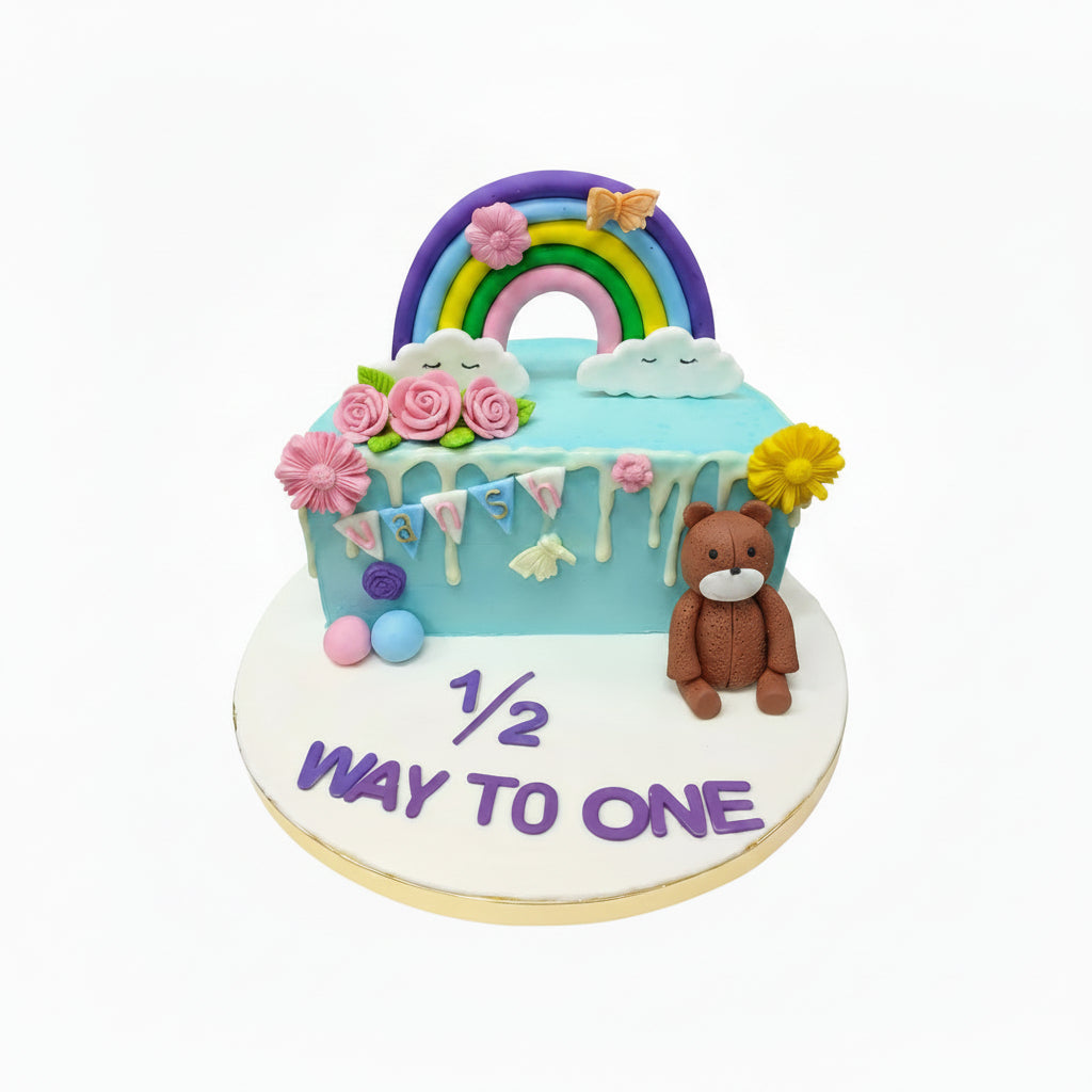 Birthday cake with rainbow, teddy bear, and '1/2 way to one' text on a white background
