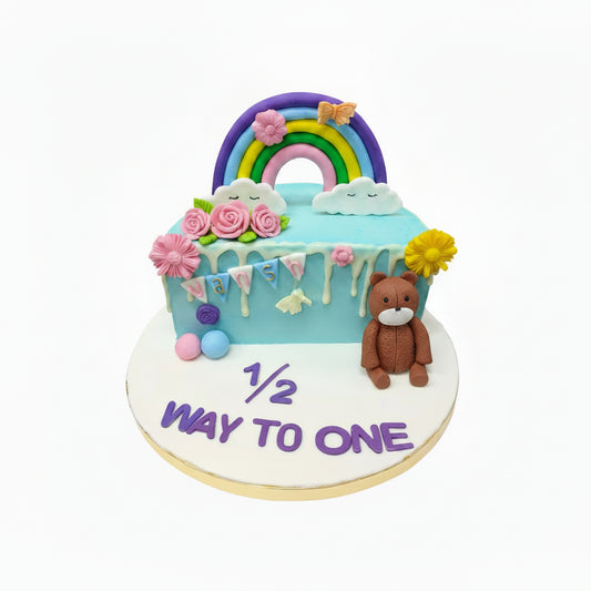 Birthday cake with rainbow, teddy bear, and '1/2 way to one' text on a white background