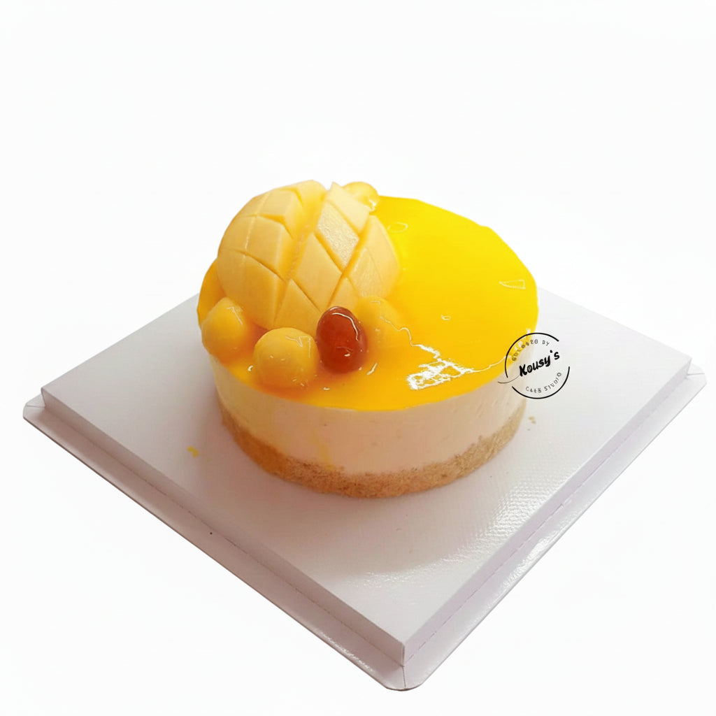 A round fruit cheesecake topped with a large mango slice, diced mango pieces, and a small red fruit, sitting on a white plate. A watermark for 'Kousy's Cake Studio' is visible on the right side.