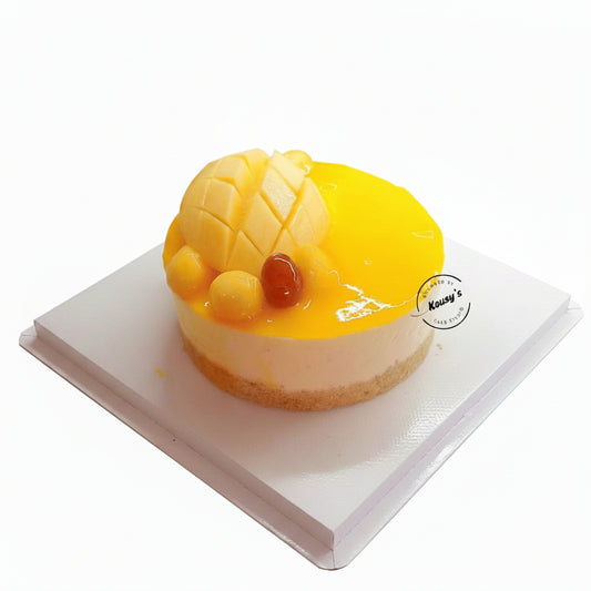 A round fruit cheesecake topped with a large mango slice, diced mango pieces, and a small red fruit, sitting on a white plate. A watermark for 'Kousy's Cake Studio' is visible on the right side.