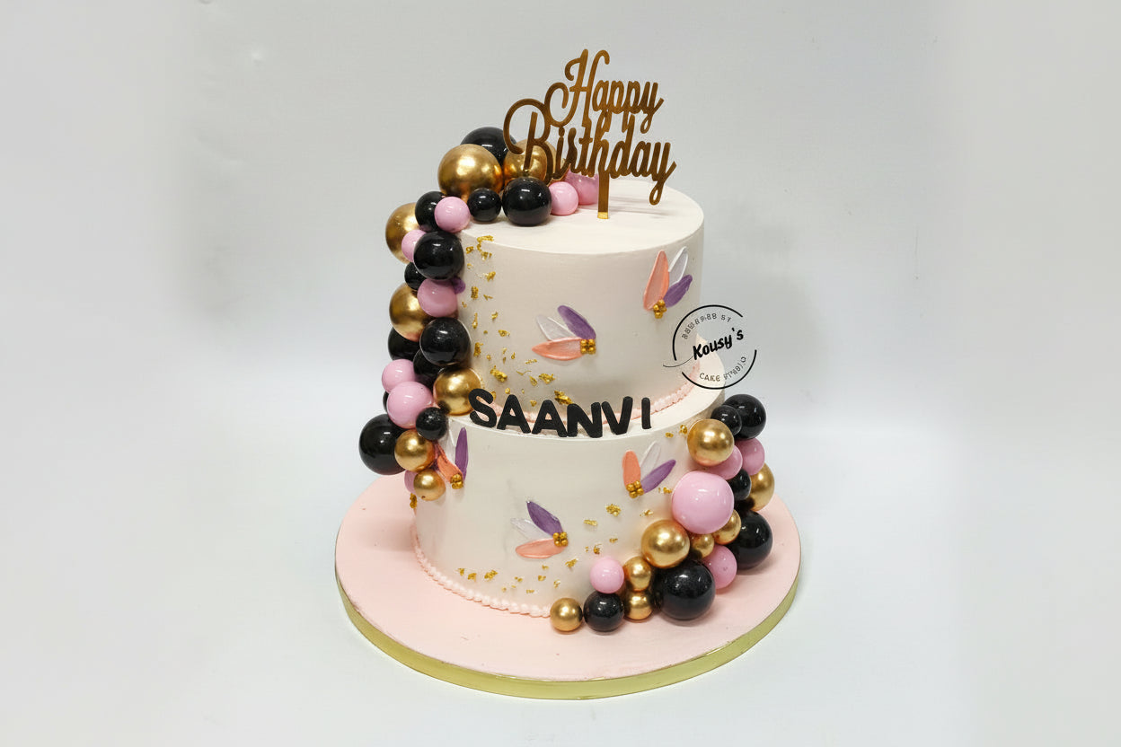 Two-tier birthday cake with gold, black and light pink faux balls, and 'Happy Birthday' topper, on a white background.
