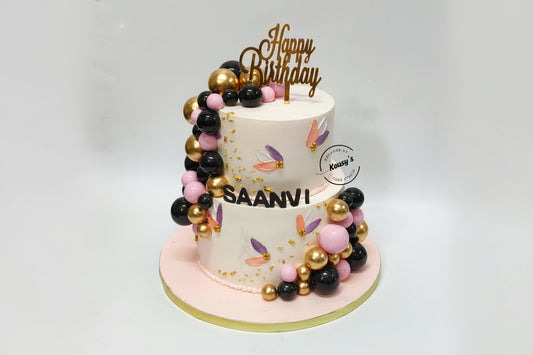 Two-tier birthday cake with gold, black and light pink faux balls, and 'Happy Birthday' topper, on a white background.
