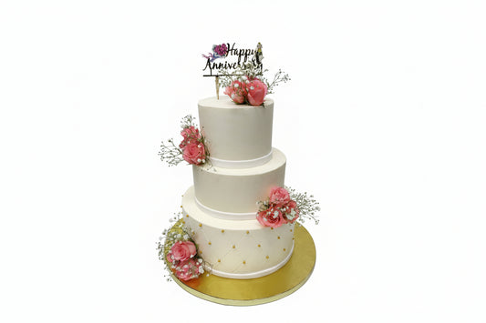 A multi-tiered white anniversary cake decorated with peach roses and a 'Happy Anniversary' topper, presented on a gold platter.